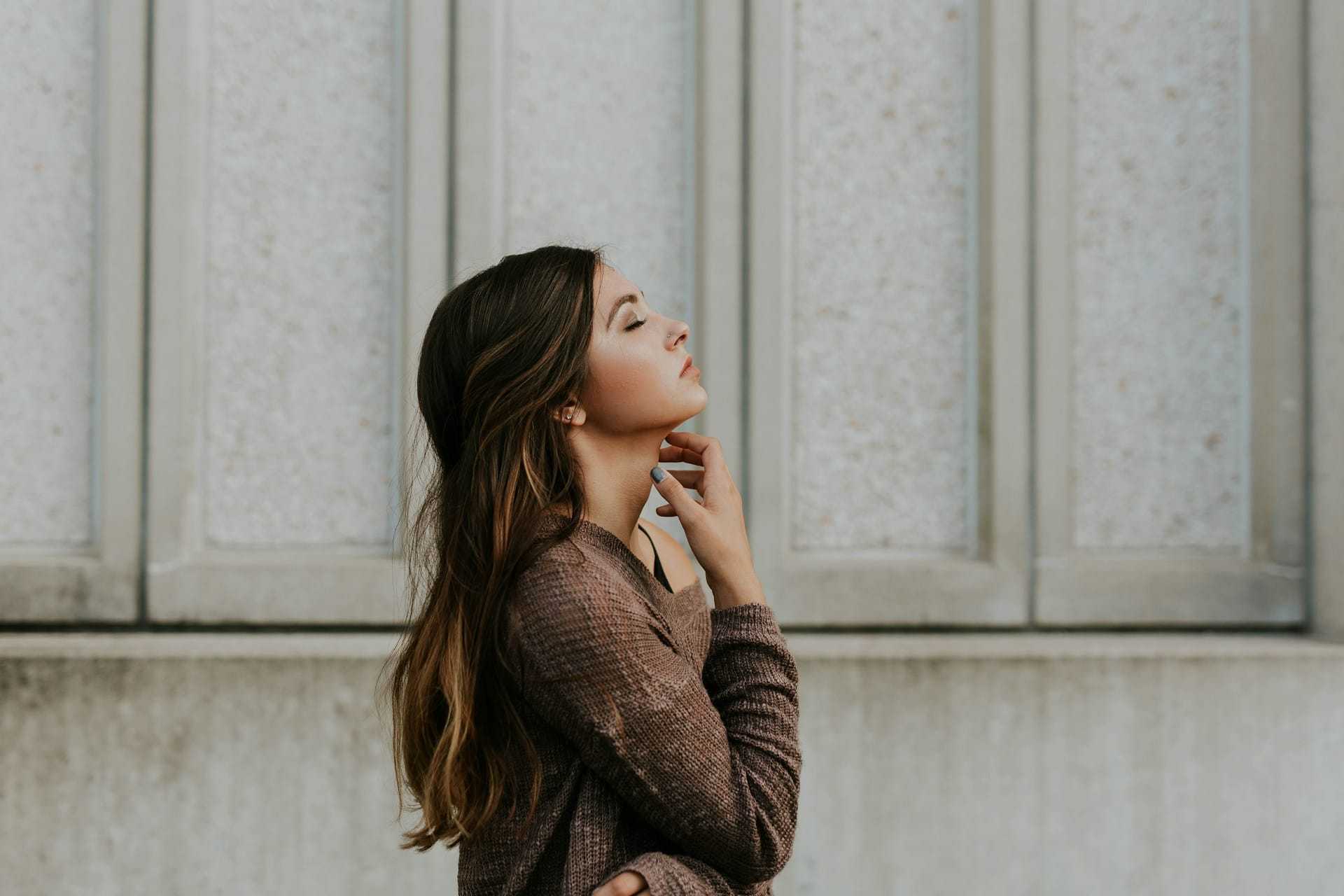 Young woman standing outdoors, looking up contemplatively against a textured wall.