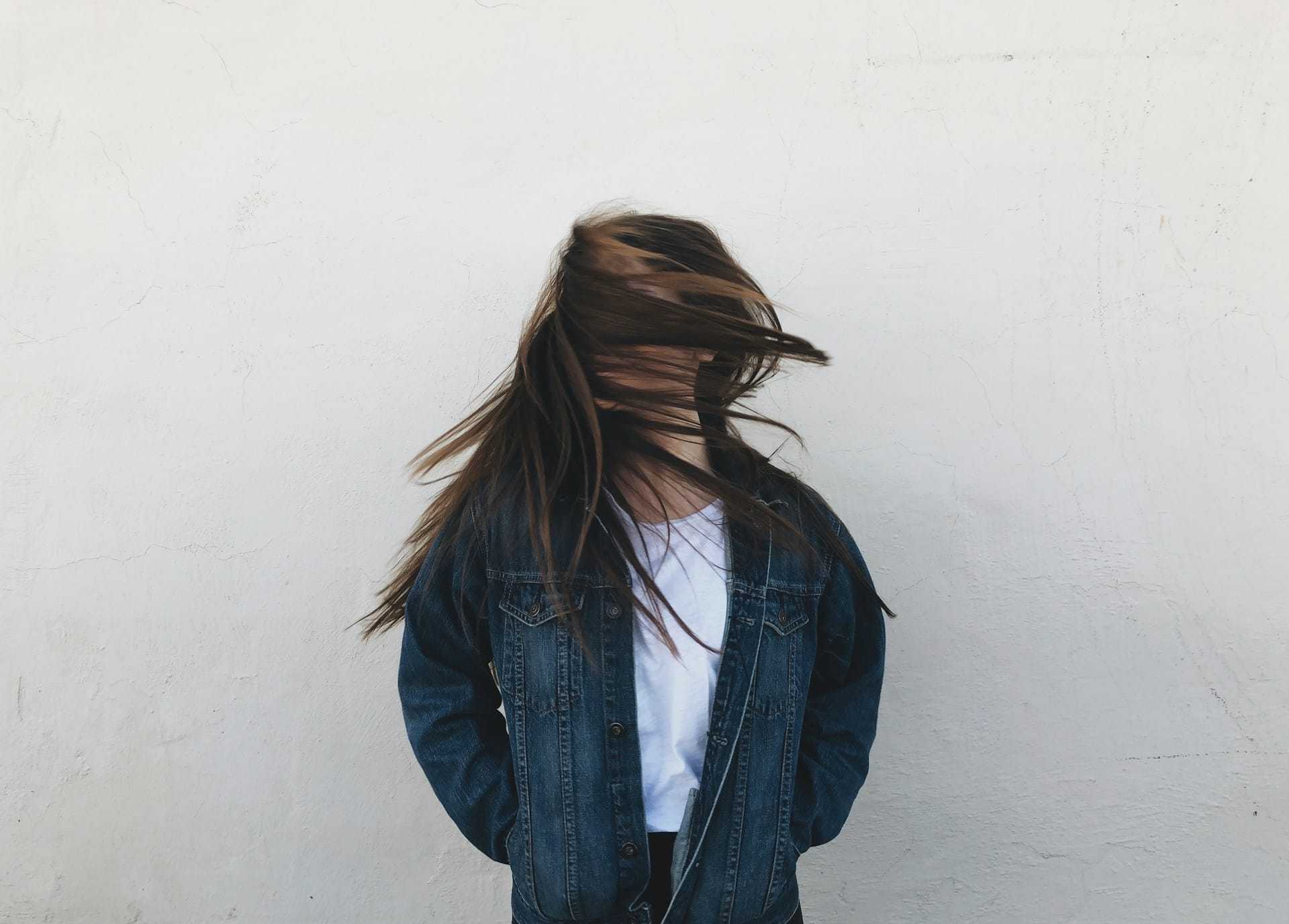 Girl in denim jacket with hair blowing over face against plain wall.