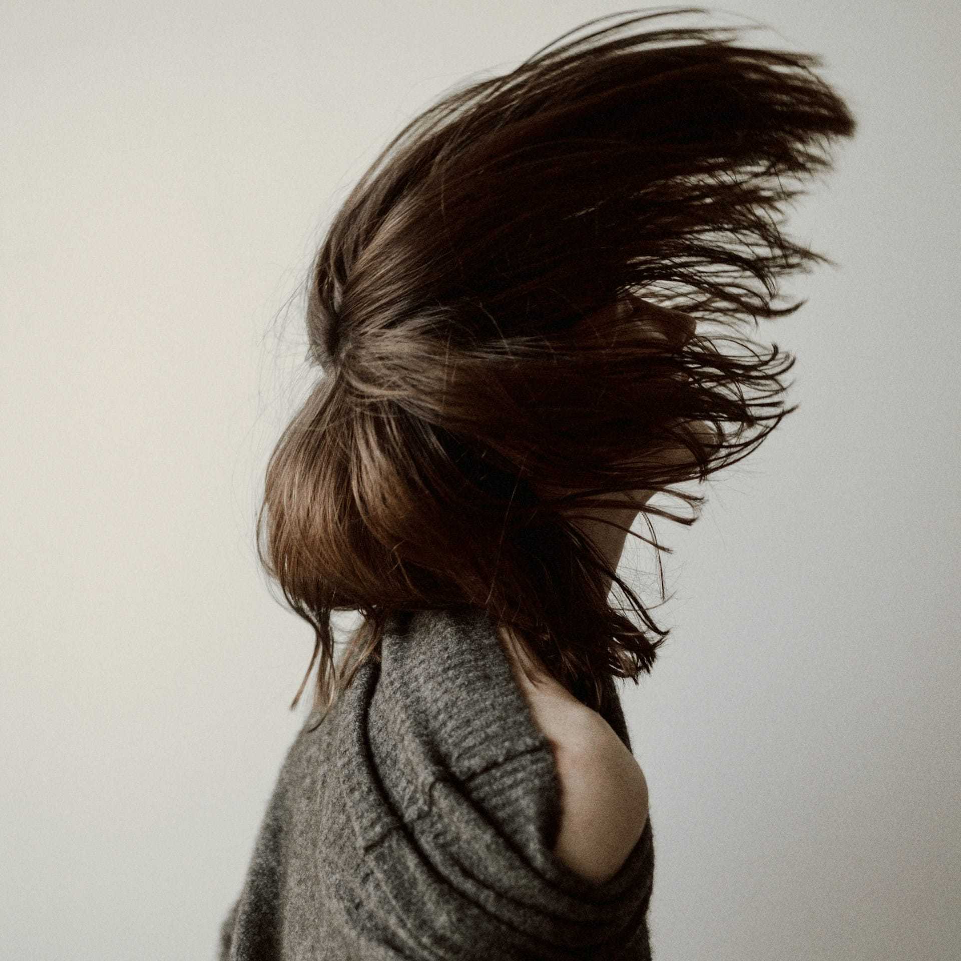 A person flipping their long hair, wearing a gray sweater with a minimalist background.